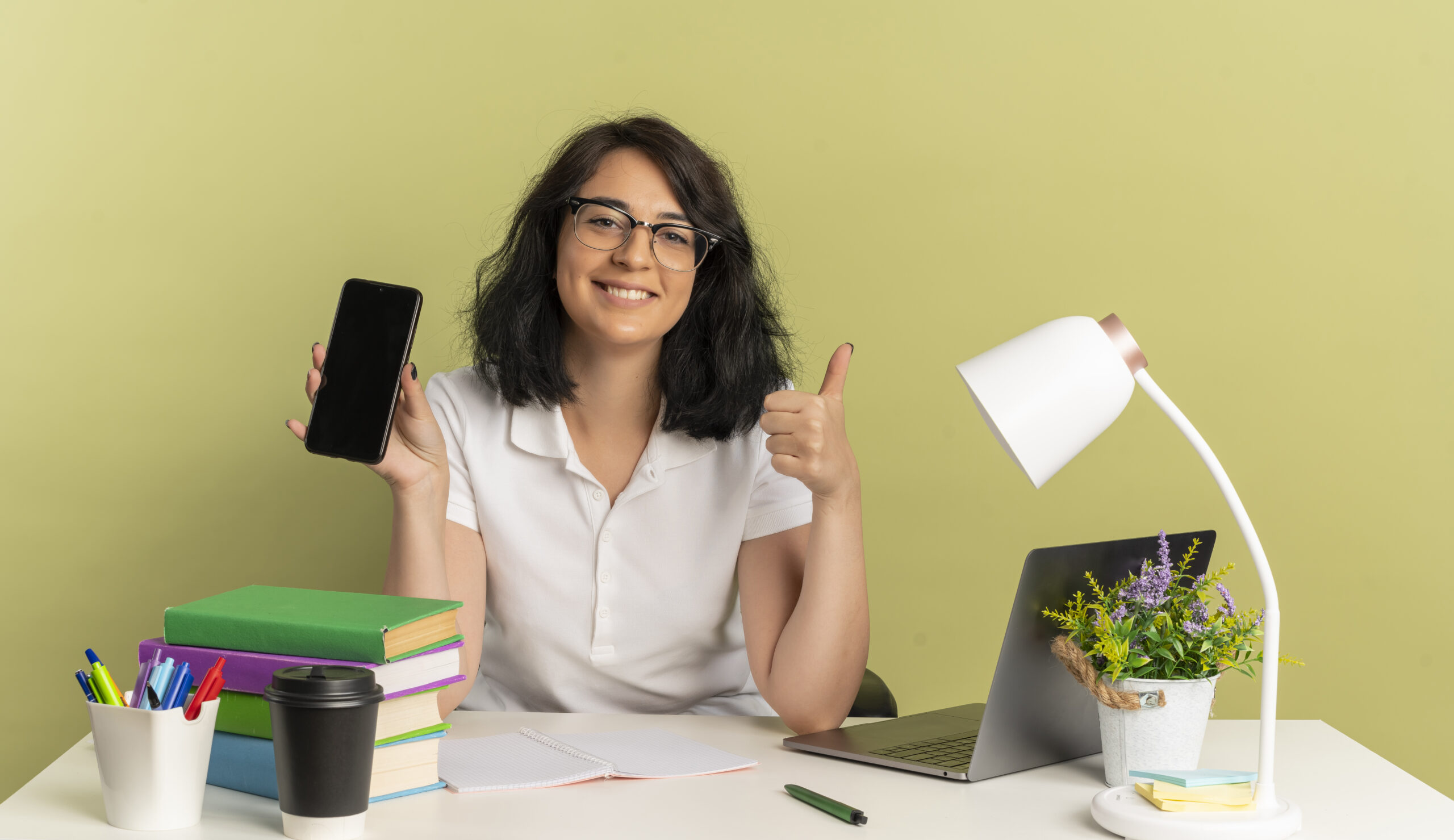 young smiling pretty caucasian schoolgirl wearing glasses sits at desk with school tools thumbs up and holds phone isolated on green background with copy space